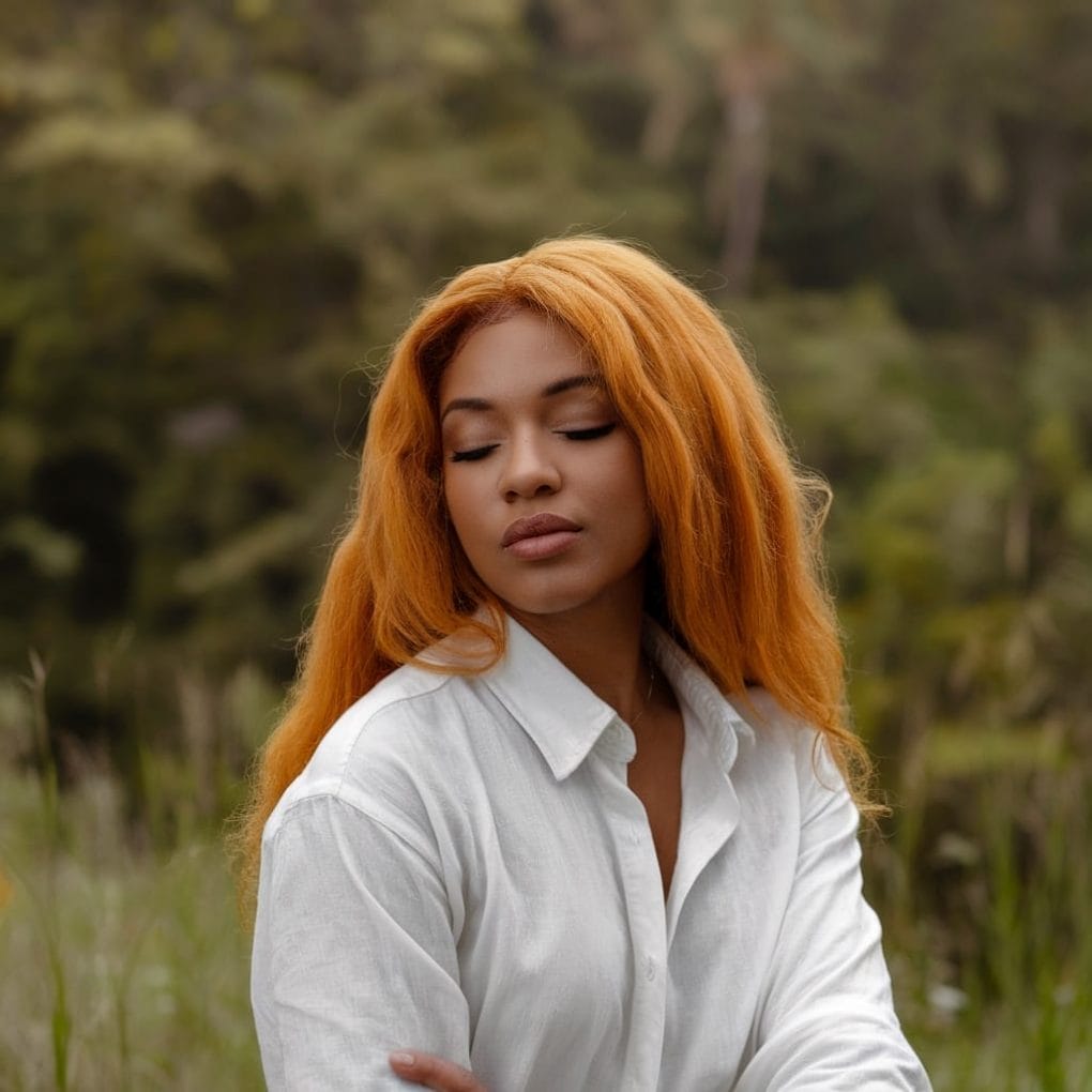 A woman with ginger hair in a white shirt, standing outdoors with a serene expression.