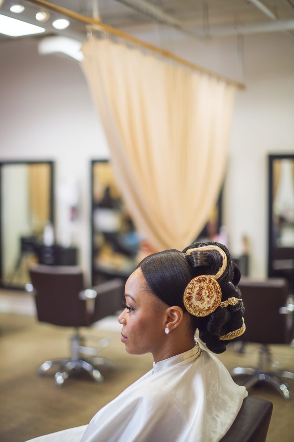 A bride with a sleek updo hairstyle adorned with a decorative hairpiece, sitting in a salon.