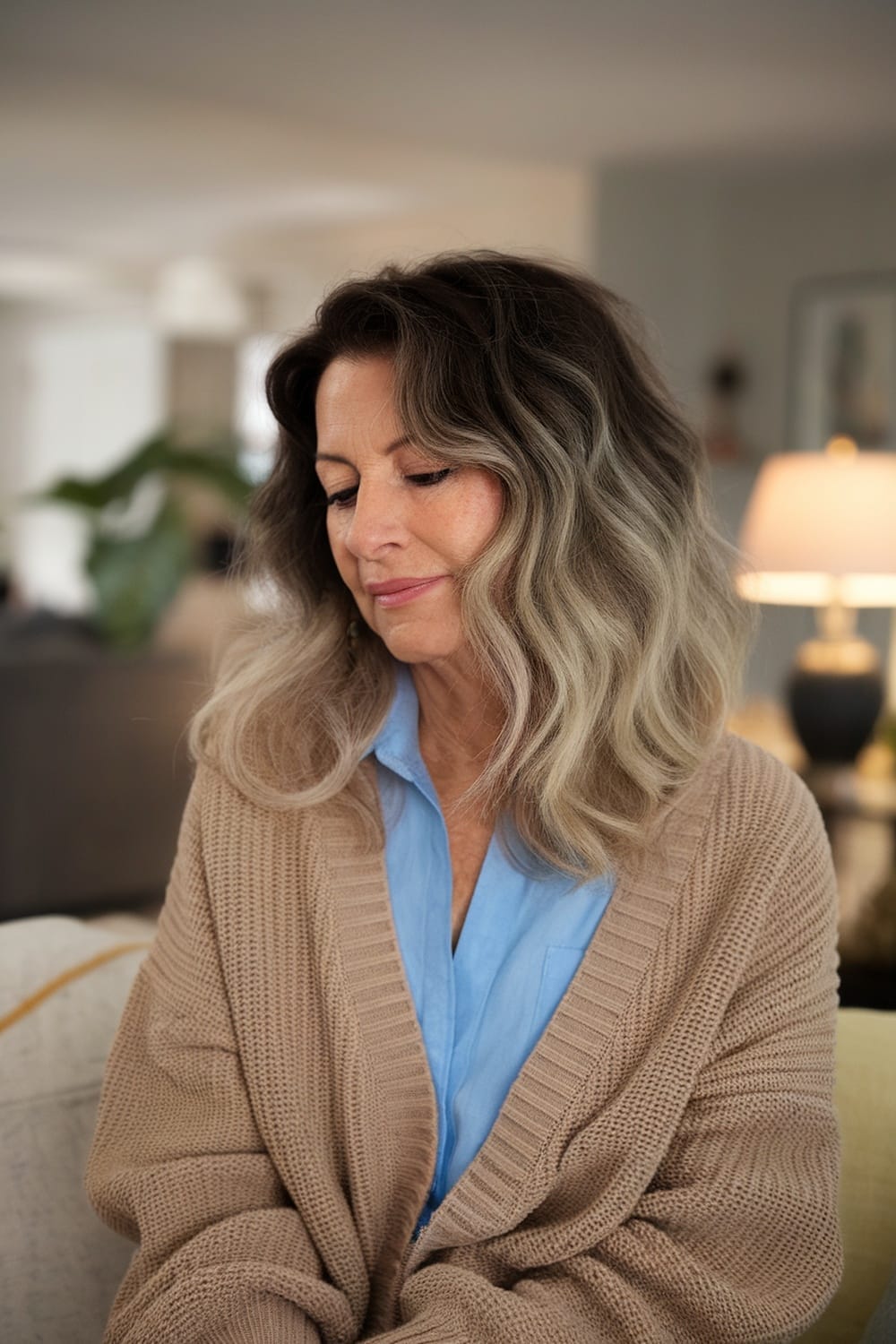 Woman with wavy hair styled in a subtle ombre, wearing a beige cardigan and blue shirt, sitting indoors.